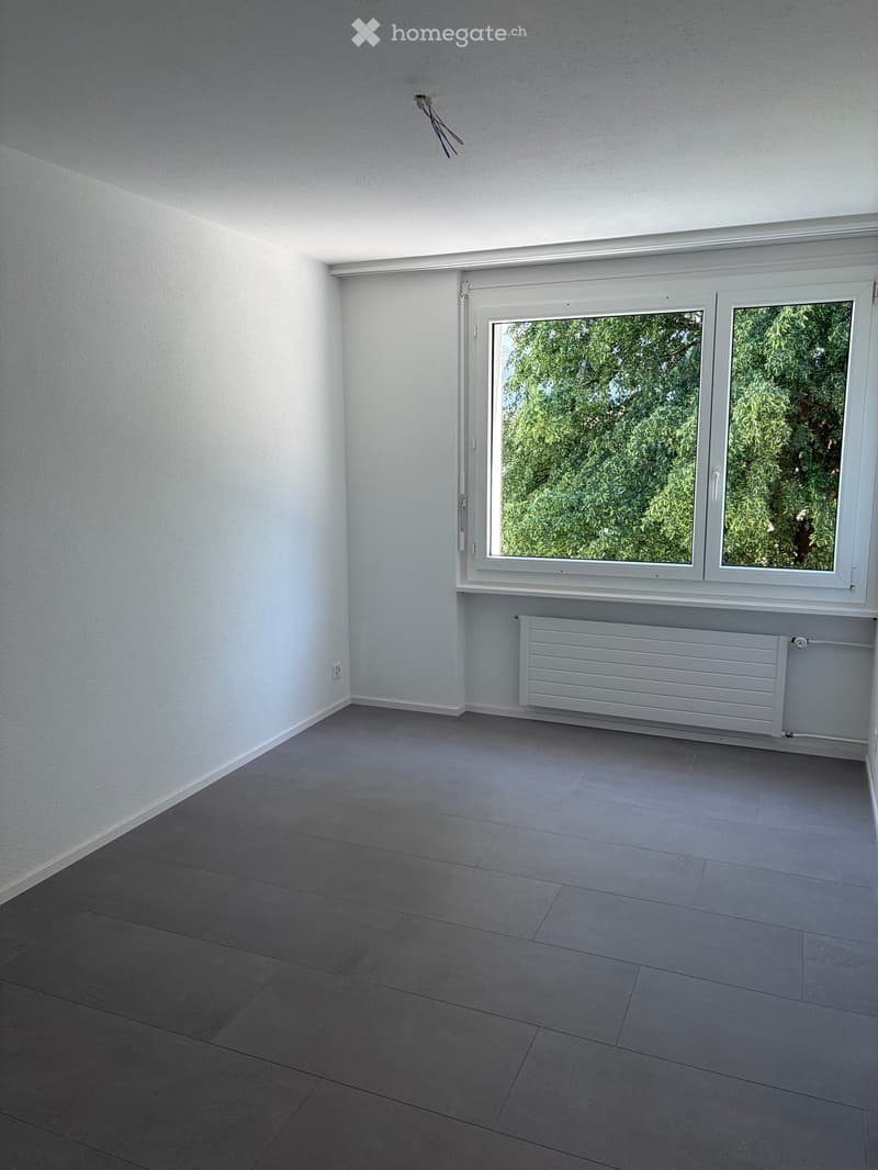 Empty room with grey tiled floor, white walls, large window with white frame, radiator below window, and electrical outlet on wall.
