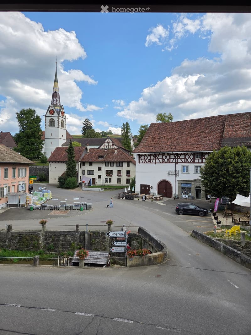 A picturesque village square with a prominent church tower, traditional houses, and a few parked cars. The area includes benches, a signpost, and some construction materials stacked on the left side.