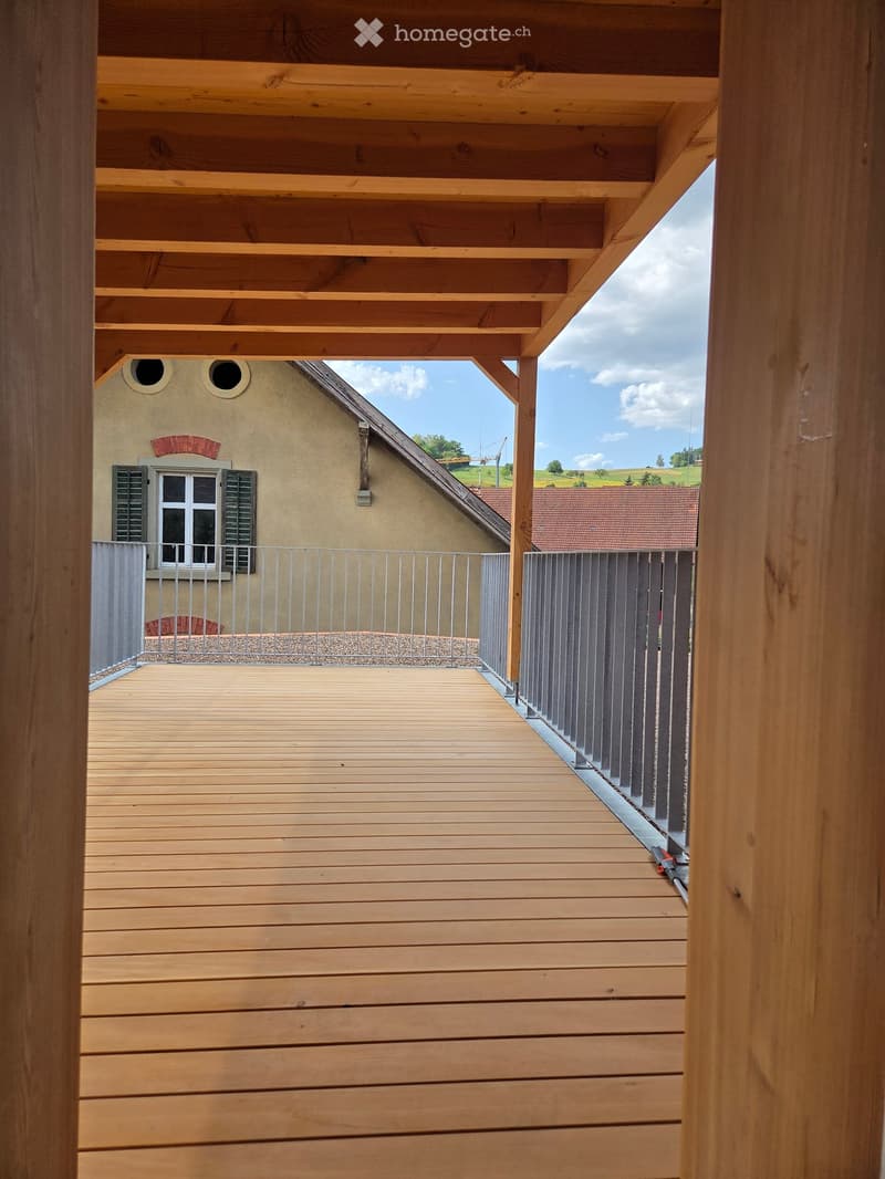 Wooden balcony with a metal railing, overlooking a house and a green landscape.