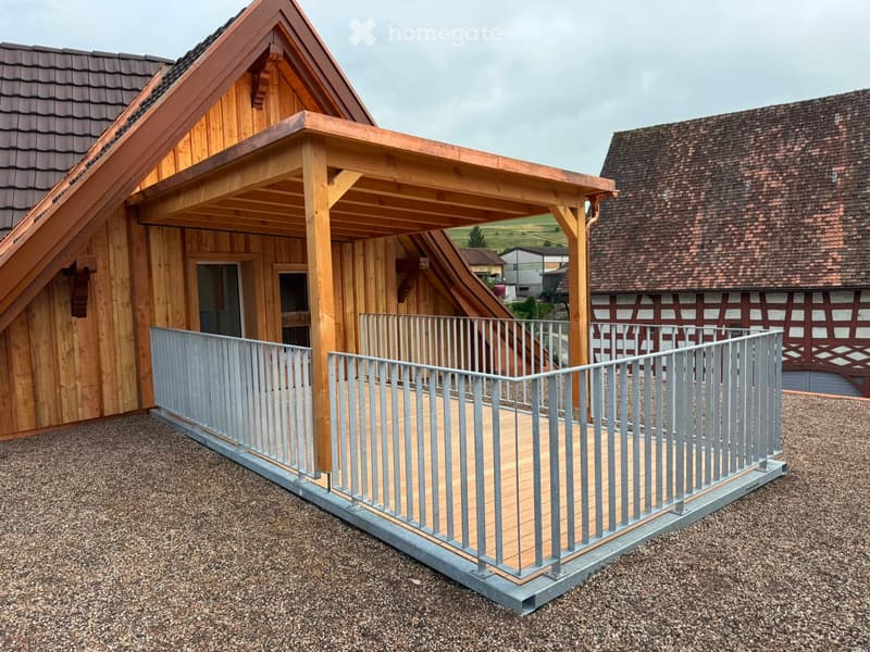Wooden balcony with metal railing, attached to a wooden house with a sloped roof and traditional half-timbered design in the background.
