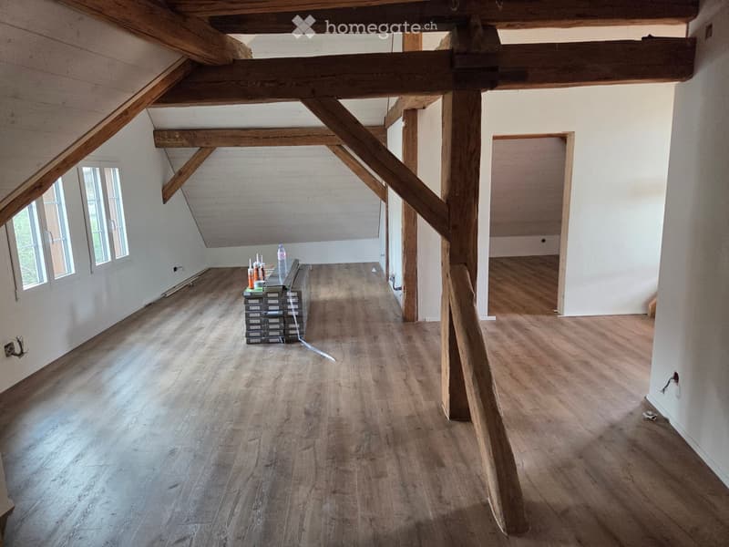 Empty room with wooden floor and exposed wooden beams, featuring a small window and a doorway leading to another room.