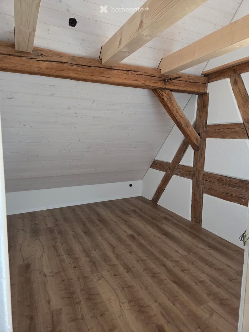 Empty attic space with exposed wooden beams, white shiplap walls, and wooden flooring.