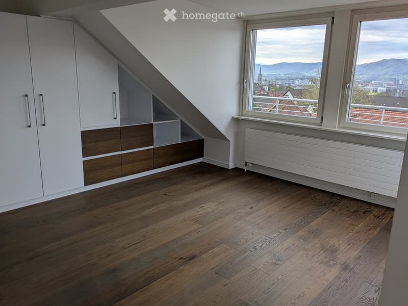 Empty attic room with wooden flooring, white walls, built-in wooden and white storage units, large window with city view, radiator, and access to a balcony.