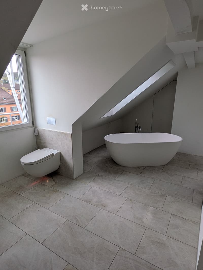 Modern bathroom with a freestanding bathtub, toilet, and large windows, featuring light-colored tiles and sleek design.