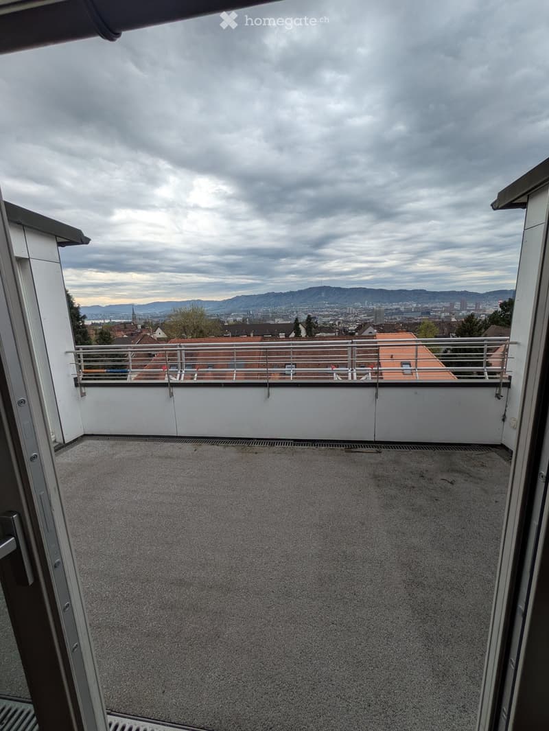 Empty balcony with a metal railing, overlooking a cityscape with buildings and distant mountains under a cloudy sky.