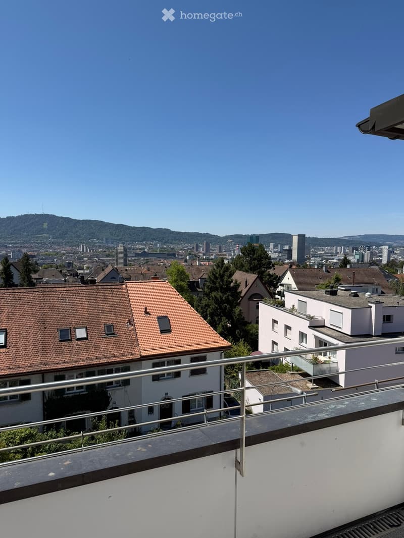 Balcony overlooking a cityscape with residential buildings, trees, and distant hills under a clear blue sky.