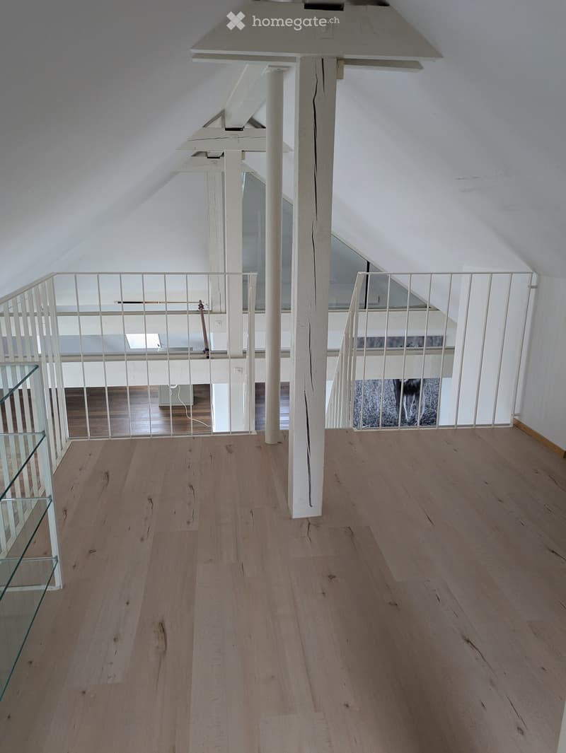 Empty room with wooden flooring, white walls, and a white railing leading to another room. The ceiling has exposed beams and skylights.