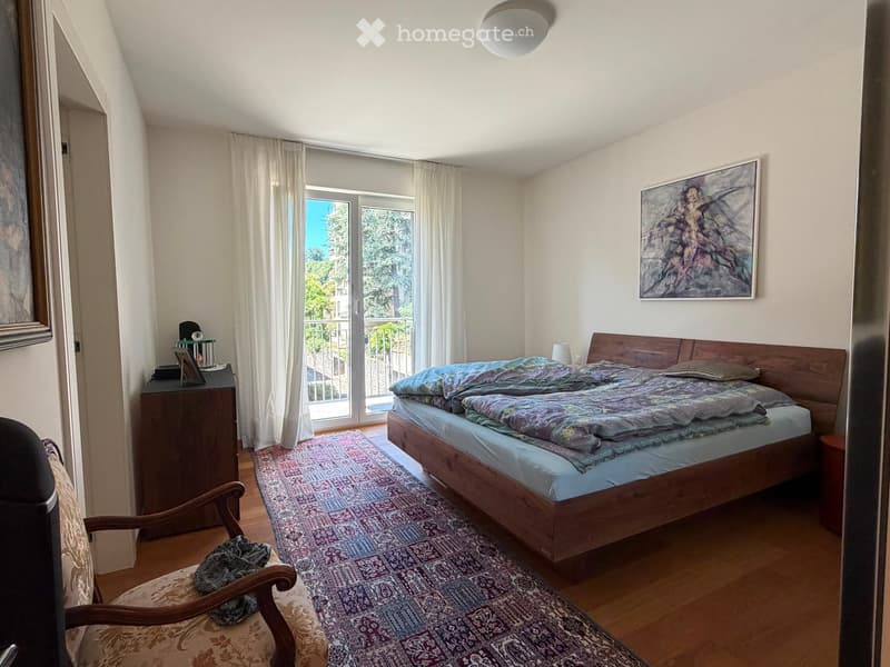 Cozy bedroom with wooden bed, patterned bedding, wooden chair, nightstand, and large window with balcony access.