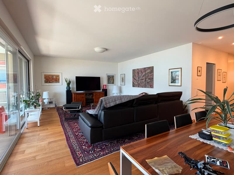 Modern living room with wooden flooring, large windows, a grey sectional sofa, coffee table, dining table, chairs, TV, and decorative plants.