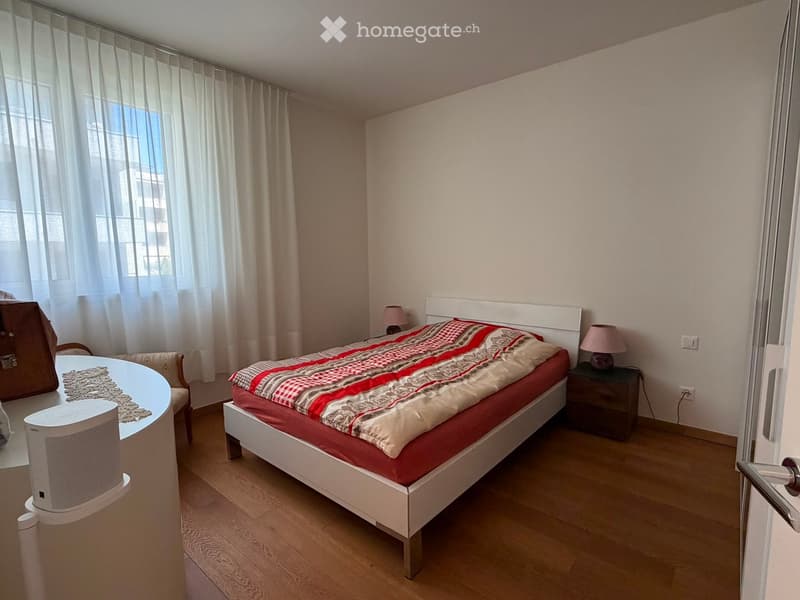 Cozy bedroom with a white bed frame, red and beige bedding, wooden nightstand with lamp, small round table with chair, and a speaker on the floor.