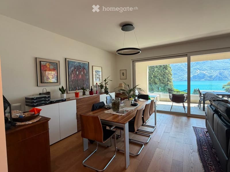 Modern living and dining area with wooden flooring, large windows offering a scenic view, a sofa, dining table with chairs, cabinet, potted plants, and artwork on the walls.