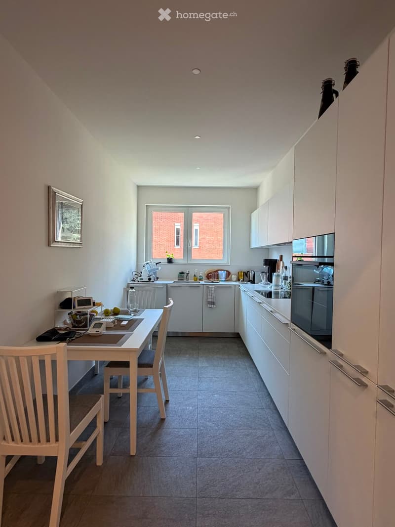 Modern kitchen with white cabinets, grey tiled floor, and a dining area with a table and chairs. The kitchen features a sink, oven, and various appliances on the counter.