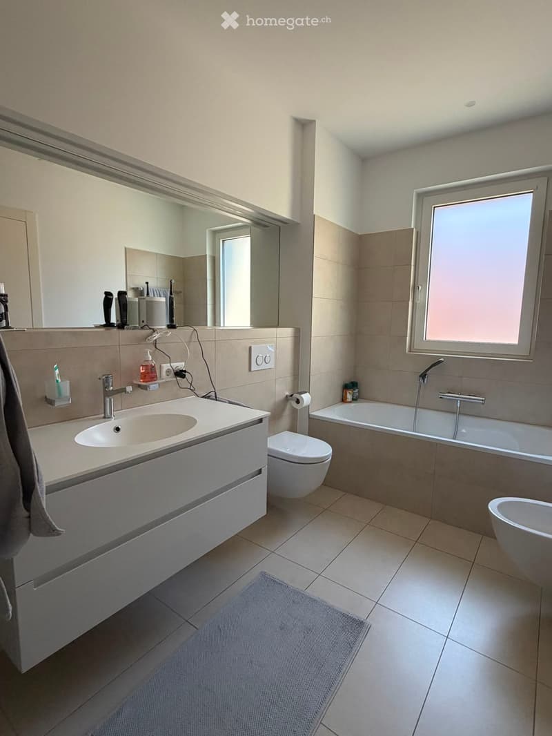 Modern bathroom with a white sink, toilet, bathtub, and large mirror. Includes a towel, toothbrush holder, soap dispenser, and electric toothbrush on the counter.