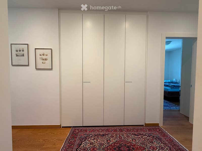 Empty room with wooden flooring, large white built-in wardrobes, and two framed pictures on the wall. An open doorway leads to another room with a bed visible.