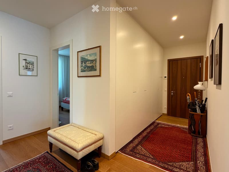 Hallway with wooden flooring, white walls, framed artwork, a small bench, and an umbrella stand. A bedroom is visible through an open doorway.
