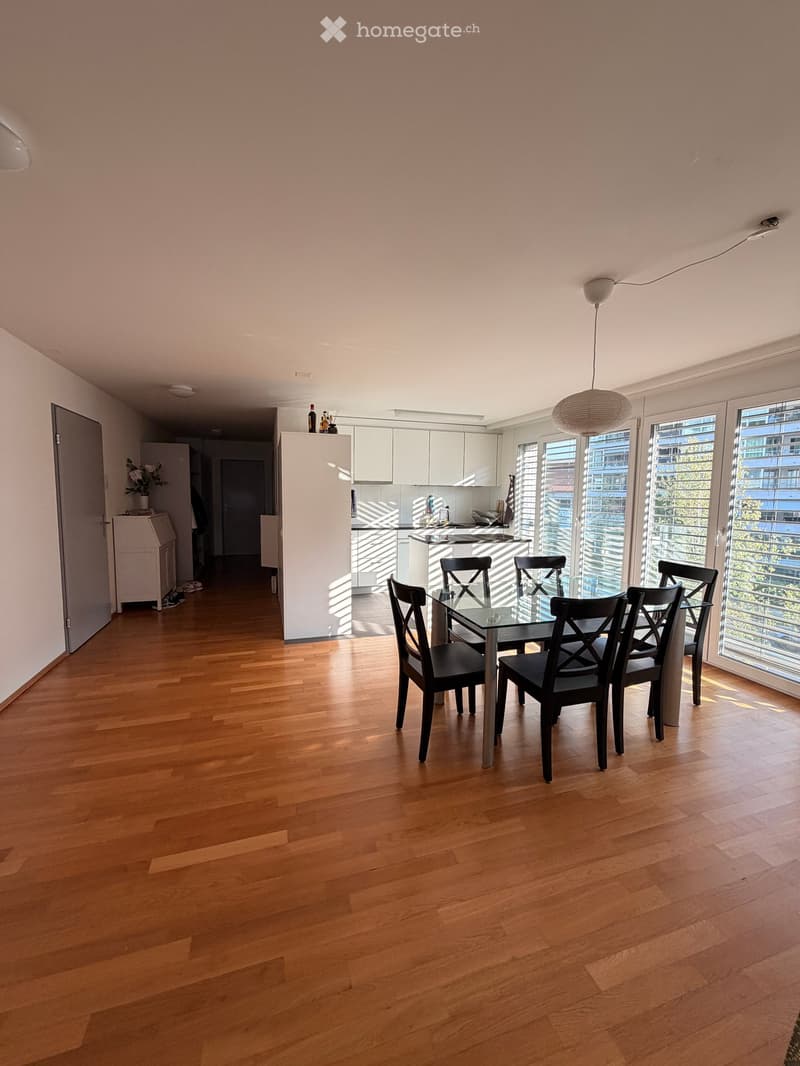 Bright dining area with wooden flooring, a glass dining table surrounded by black chairs, and a modern kitchen area with white cabinets and stainless steel appliances.