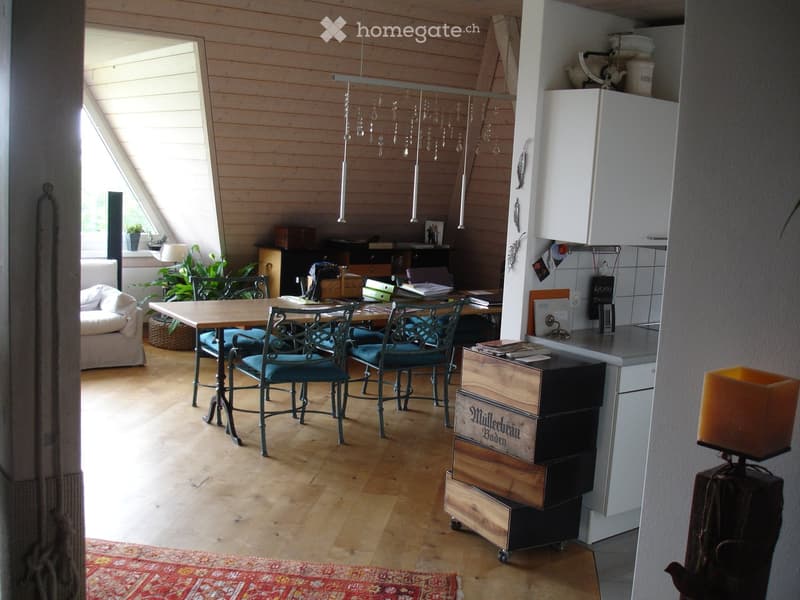 Bright, open room with wooden floors, a dining table with chairs, a sofa, a kitchen area with cabinets, and a hanging light fixture.