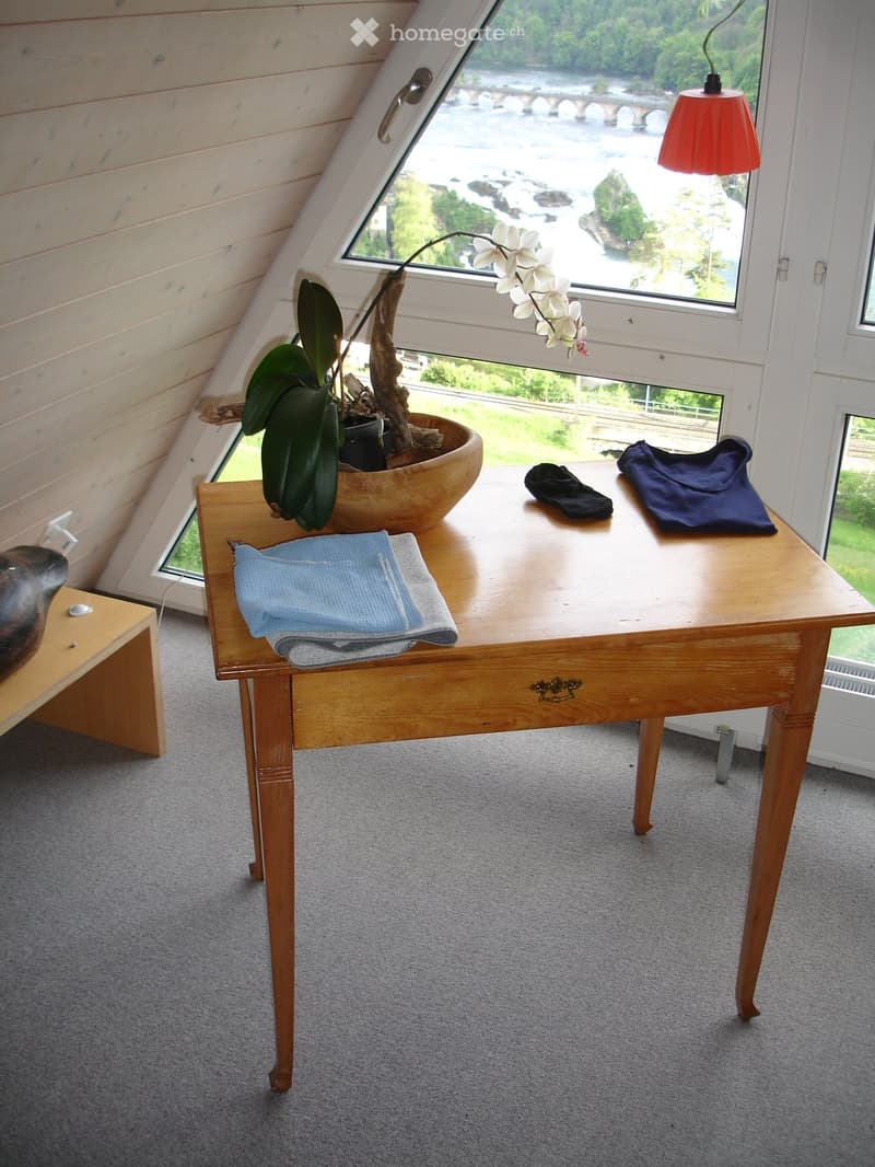 A small, cozy room with a wooden table, a stool, a potted plant, and towels on the table. The room has large windows offering a scenic view of a river and landscape.