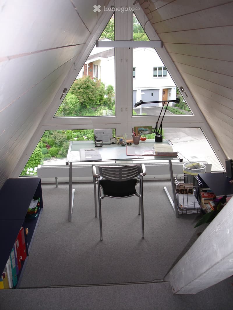 Bright attic room with slanted wooden ceilings, large windows, a glass desk, a modern chair, a lamp, and a shelf with books and magazines.