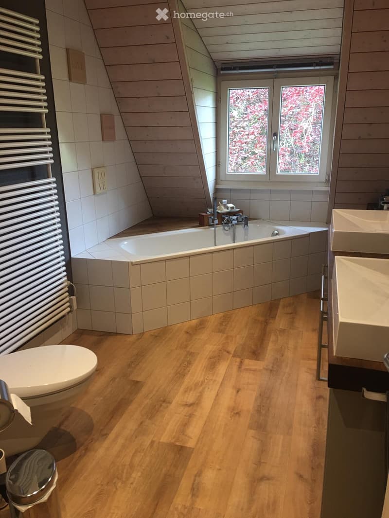 Modern bathroom with wooden flooring, a bathtub, toilet, sink, and a radiator. The room features large windows with a view of greenery.