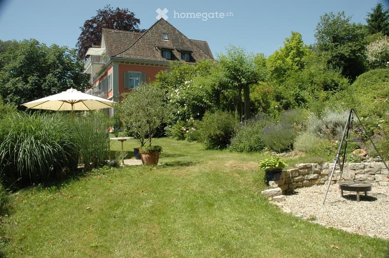Well-maintained garden with a lawn, various plants, a stone wall, a patio umbrella, and a small table. A two-story house with a red facade and a balcony is visible in the background.