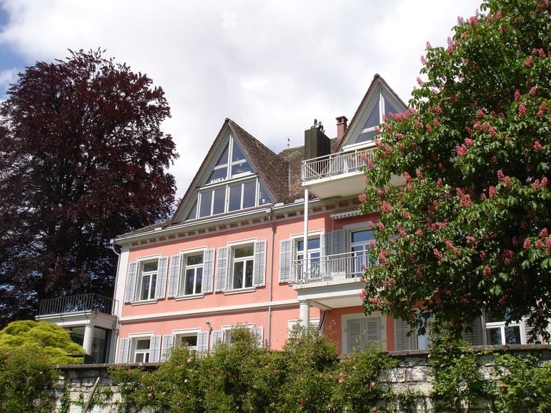 Two-story pink house with white trim, multiple windows, balconies, and a triangular roof section.