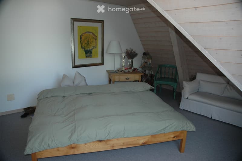 Cozy attic bedroom with a wooden bed, grey duvet, nightstand with lamp, small sofa, green chair, and decorative items on a small table.