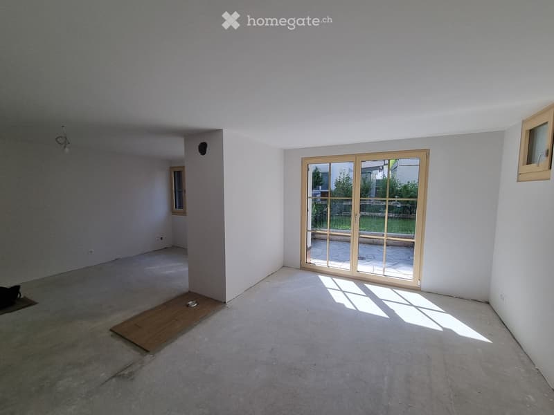 Empty room with white walls, concrete floor, large sliding glass door leading to outdoor area, small window, and a wooden plank on the floor.