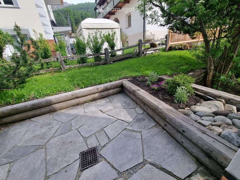 Garden area with stone-paved patio, wooden log border, small garden bed with plants, and a wooden fence surrounding the space.