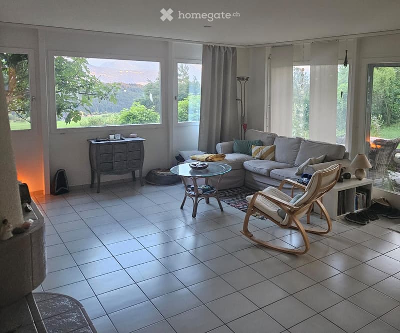 Bright living room with large windows, a grey sectional sofa, a glass coffee table, a wooden rocking chair, and a side table with a lamp.