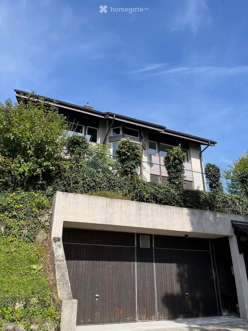 Modern two-story house with a raised ground floor, large garage, and balcony on the upper level, surrounded by greenery.
