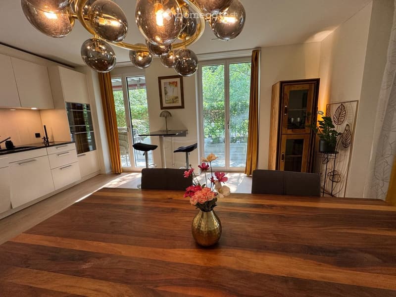 Open-plan kitchen and dining area with modern white cabinets, a wooden dining table, and a vase of flowers in the center. The space features large windows with curtains, a chandelier, and a small seating area with stools.