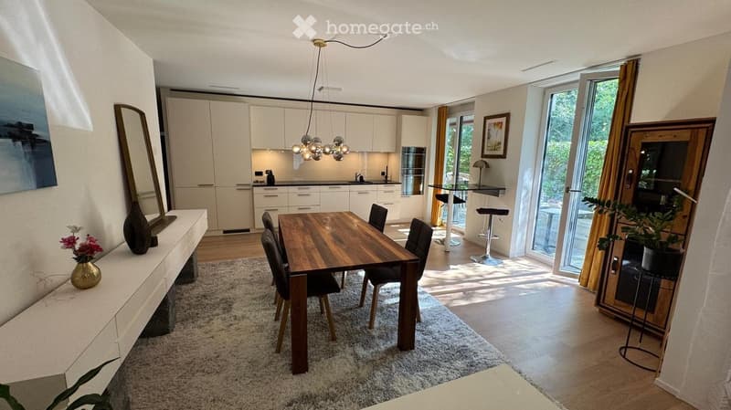 Modern open-plan living and dining area with a wooden dining table, chairs, kitchen cabinets, and a sleek kitchen sink. The space features a large mirror, decorative vases, and a potted plant, with natural light streaming in from large windows and sliding