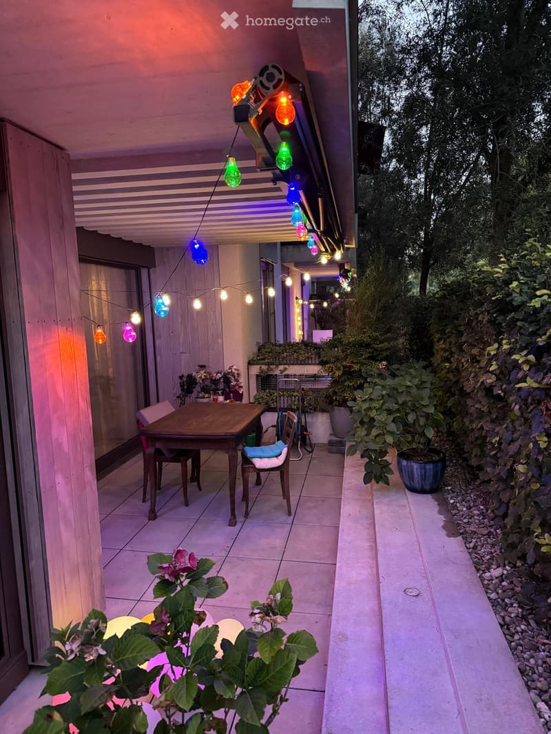 Balcony with a wooden table, chairs, colorful string lights, and potted plants.