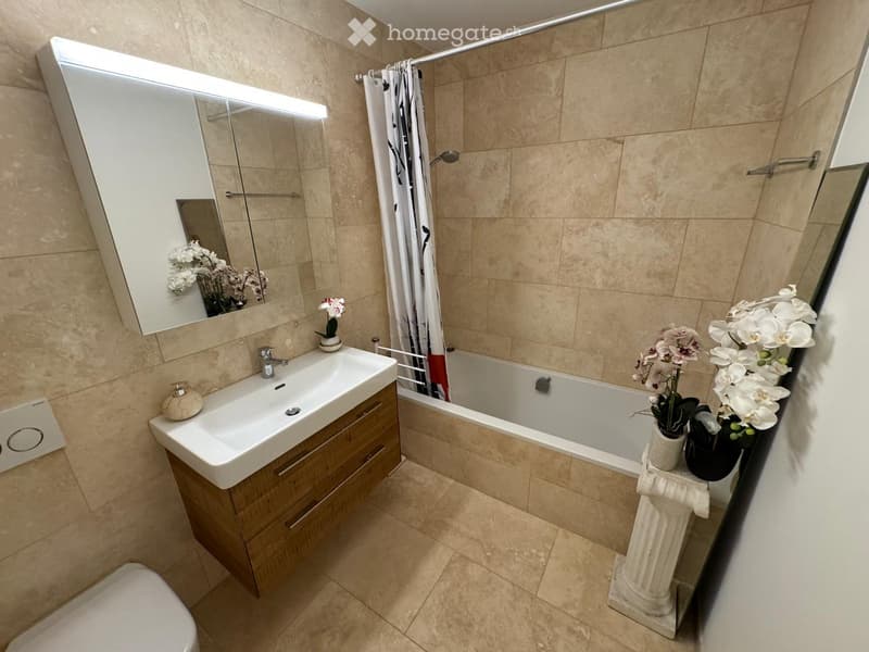 Modern bathroom with beige tiles, a wooden vanity with a sink, a bathtub with a glass shower curtain, a toilet, and decorative flower vases on a pedestal.