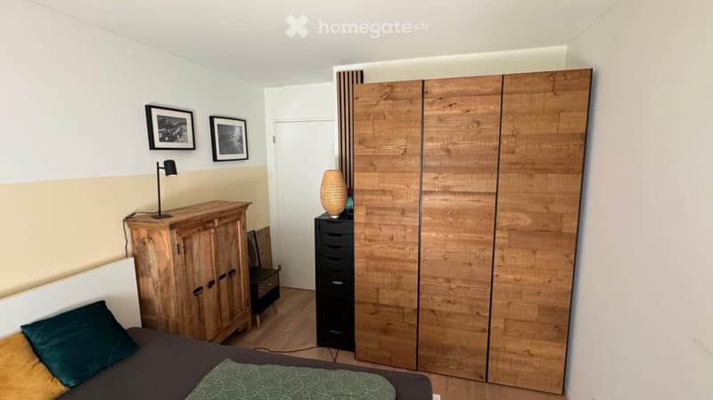 Cozy bedroom with a wooden wardrobe, a bed with pillows, a nightstand with a lamp, and a drawer unit with a decorative vase on top.