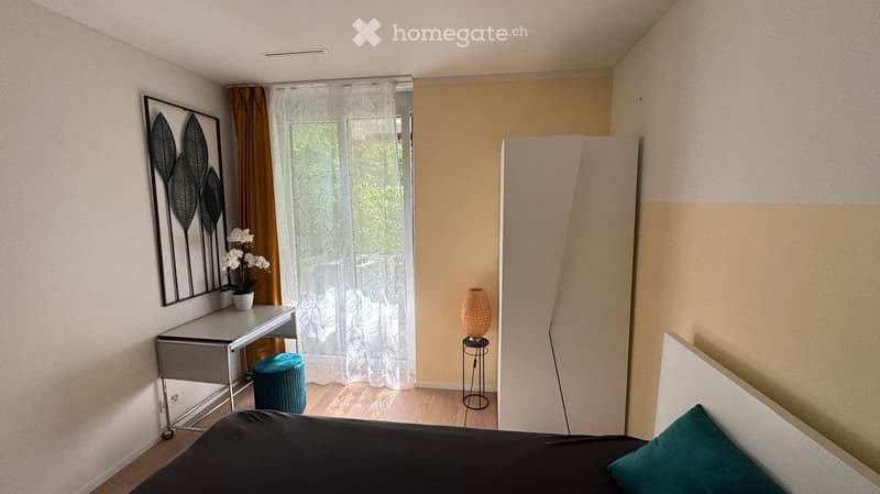 Modern bedroom with a bed, desk, stool, lamp, vase, and picture frame. The room features light-colored walls, wooden flooring, and sheer curtains on the window.
