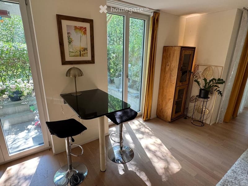 Bright room with wooden flooring, large windows, two black stools, a glass table, a lamp, a wooden cabinet, a mirror, and a potted plant.