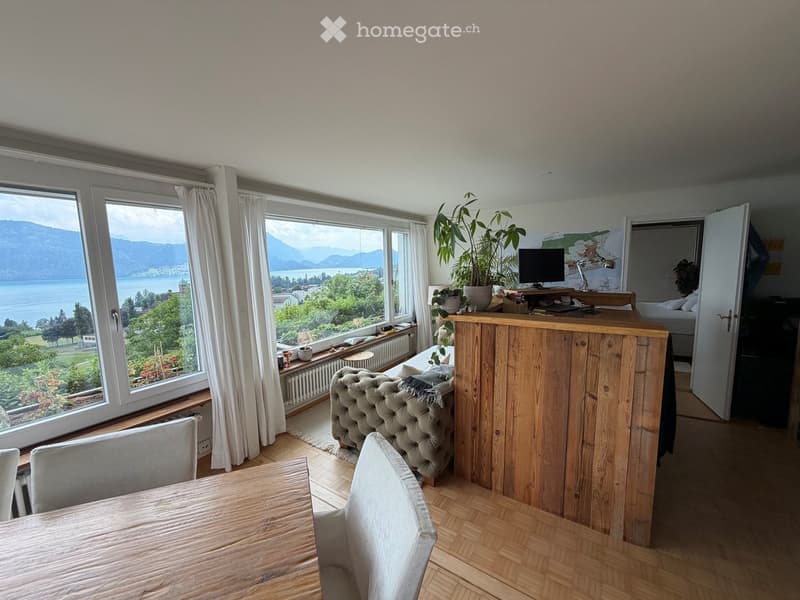 Bright living space with large windows overlooking a scenic lake and mountains, featuring a wooden table, sofa, and a small kitchen counter.