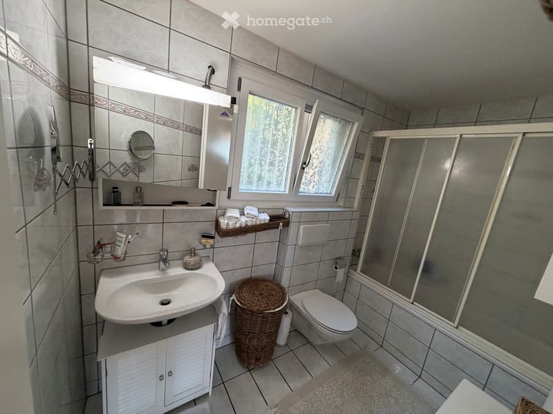 Bathroom with a sink, toilet, mirror, cabinet, wicker basket, and shower stall. The walls are tiled, and there is a window with a floral curtain.