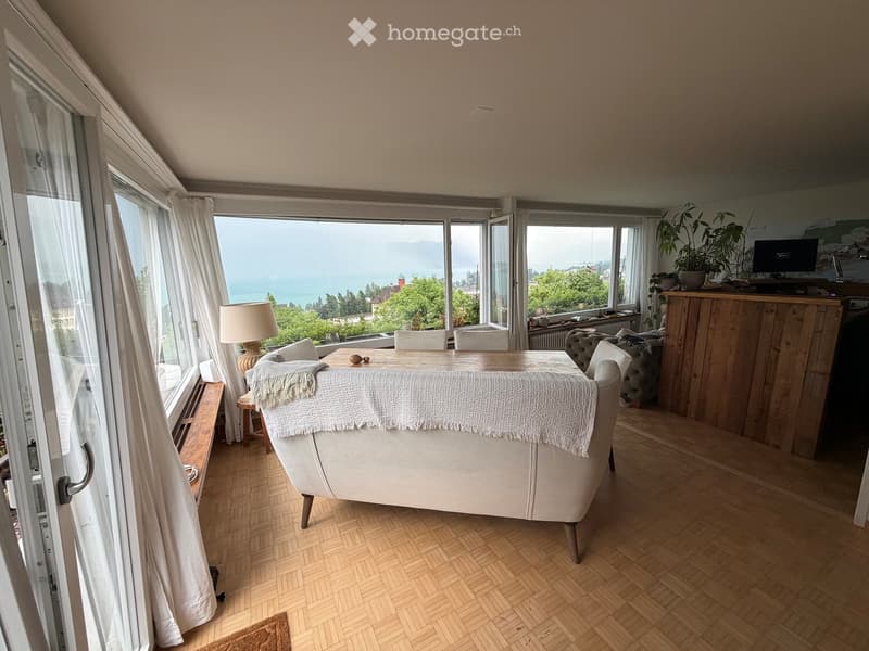 Bright living room with large windows offering a scenic view, featuring a white sofa, wooden table, chairs, a lamp, a television, a bar counter, and plants.