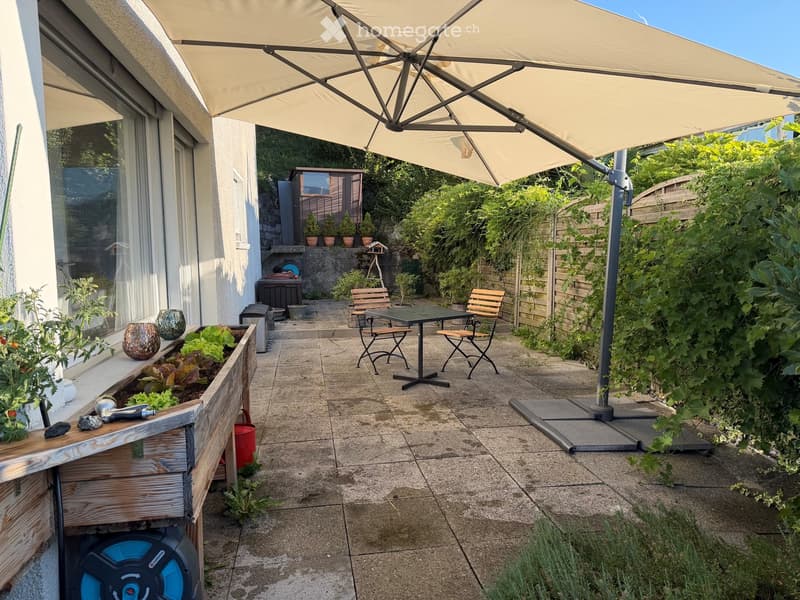 Patio area with a large umbrella, outdoor dining table and chairs, potted plants, and a raised garden bed with vegetables.