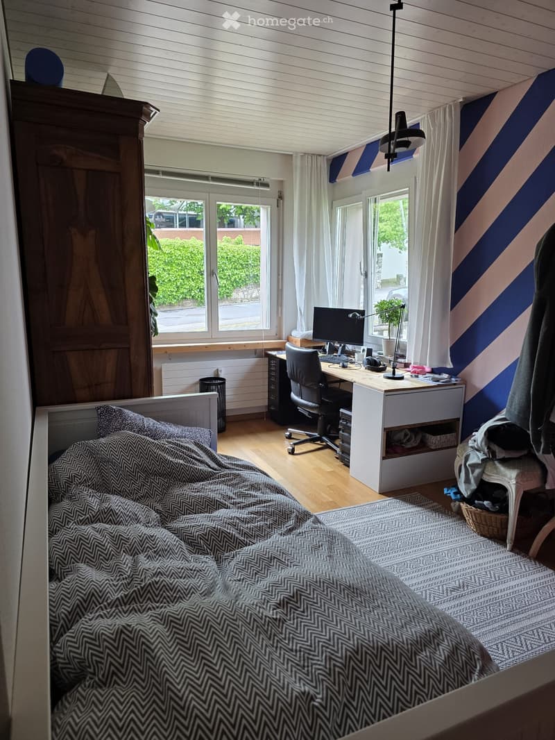 Cozy bedroom with a bed, desk, chair, and computer. Features wooden flooring, white walls with blue stripes, and large windows with white curtains.