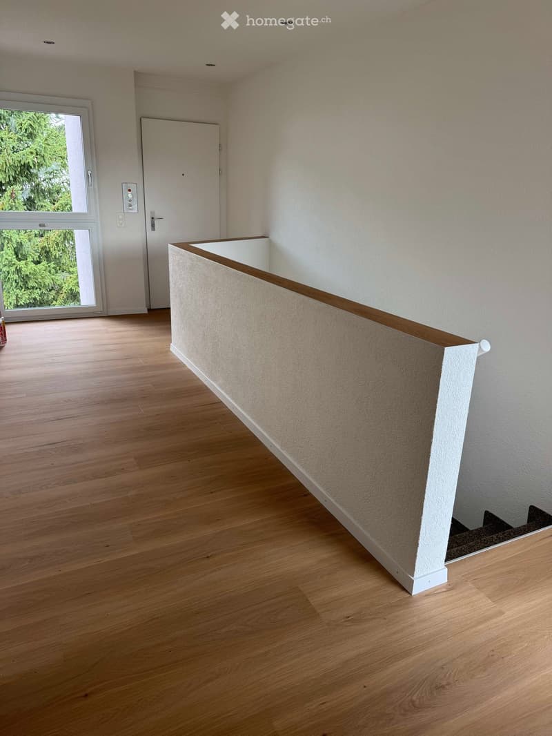 Empty room with wooden flooring, white walls, and a staircase leading upwards. A large window with a view of greenery is visible on the left side.