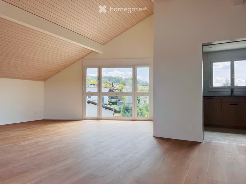 Empty room with wooden flooring, white walls, large windows, and a slanted wooden ceiling.