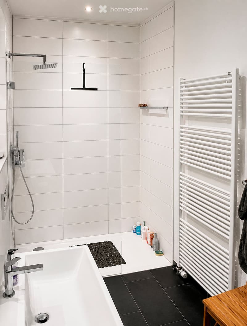 Modern bathroom with a white bathtub, chrome faucet, showerhead, radiator, and various toiletries on the shelf.