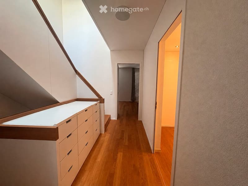 Modern hallway with wooden flooring, white walls, built-in storage unit with drawers, and a skylight providing natural light. The hallway leads to another room with a visible door and window.