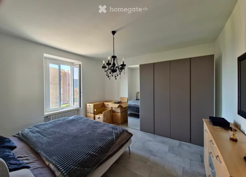 Modern bedroom with a large window, a bed with striped bedding, a wardrobe, a dresser, and a chandelier. Some boxes are visible, indicating the room is being unpacked or organized.