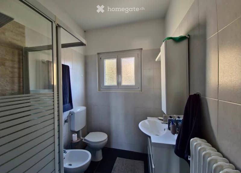 Modern bathroom with a toilet, sink, mirror, shower, radiator, and towel. The space features grey tiles and a window for natural light.