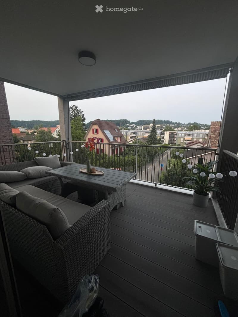 Balcony with wooden decking, metal railing, sofa, table, vase, and potted plant, overlooking a town with houses and trees.